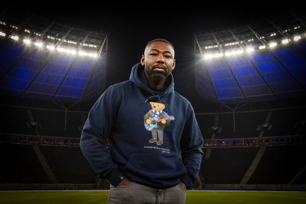 Mann steht im beleuchteten Olympiastadion und trägt den dunkelblauen Frank Zander Charity-Hoodie. Das Brustmotiv zeigt einen coolen Bären mit Sonnenbrille, blau-weißem Schal und einer Gitarre.