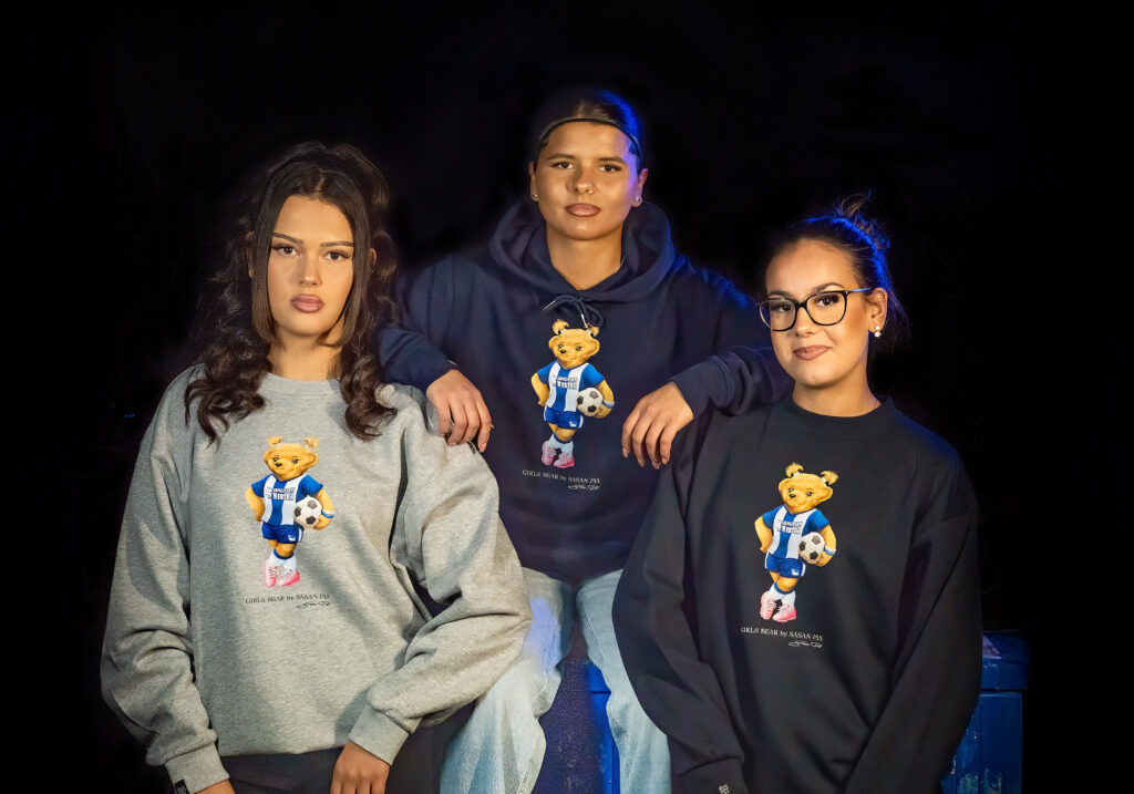 Drei weibliche Fans präsentieren die neue Girls Bär Kollektion. Sie tragen graue und dunkelblaue Sweater und Hoodies mit dem weiblichen Hertha-Bären-Motiv, der ein Trikot trägt und einen Fußball hält.